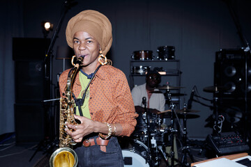 Portrait of young musician playing saxophone during band rehearsal in music studio, wearing stylish...