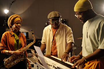Group of three musicians engaged in music performance in studio setting, with one playing saxophone and another on keyboard while third member observes attentively