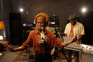 African American woman passionately singing into a microphone with band members playing instruments in recording studio atmosphere