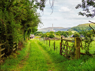 A serene country pathway in a lush, green landscape with distant hills and industrial chimneys on the horizon.