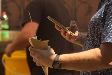Midsection of man holding cone with french fries in one hand, smartphone in the other hand and...