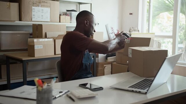 Medium shot of black male manager checking parcels in boxes with scanner while working at desk with laptop in storage office