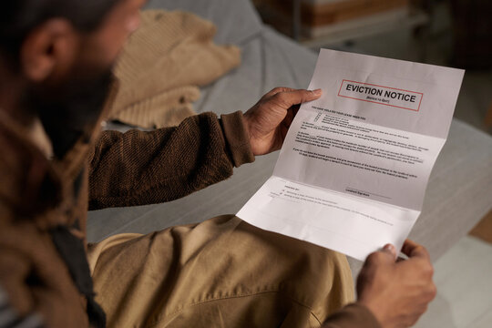Person holding eviction notice while sitting on couch and reading in a dimly lit room without natural light coming in through the window behind