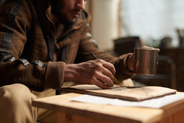 Person sitting at wooden table, writing notes in notebook while holding metal cup in cozy home environment with warm ambient lighting