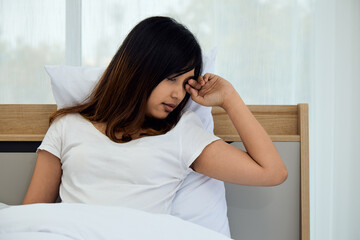 Young woman sitting up in bed, rubbing her eyes with sleepy expression. She is leaning against headboard, wrapped in white blanket in bright, airy bedroom