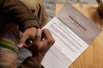 Person sitting on comfortable chair and reading eviction notice placed on wooden table. Hands and...