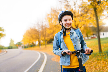 Happy kid riding scooter on autumn road to school. Teenager in protective helmet on cycleway. Girl smiling on town street. Safety childhood, healthy outdoor activities. Authentic lifestyle moment