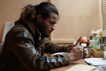 Man deeply engrossed in his work while sitting at table in warmly lit setting with various items...