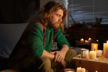Man with long hair sitting by candlelight in cozy room reflecting on thoughts, wearing green...