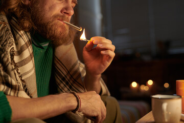 Man lighting cigarette with candle in dim cozy room, illuminating his face. He wearing warm knitted...