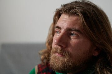 Man with long hair and beard sitting indoors, looking away thoughtfully into distance. Soft natural...