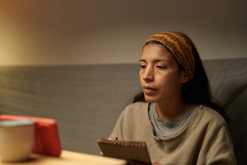 Hispanic woman sitting indoors with thoughtful expression, holding notebook and pen, appearing in...