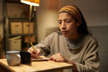 Contemplating woman sitting at wooden table, writing notes in notepad with pencil in cozy living room setting, lamp providing soft illumination, wearing colorful headband, focused expression