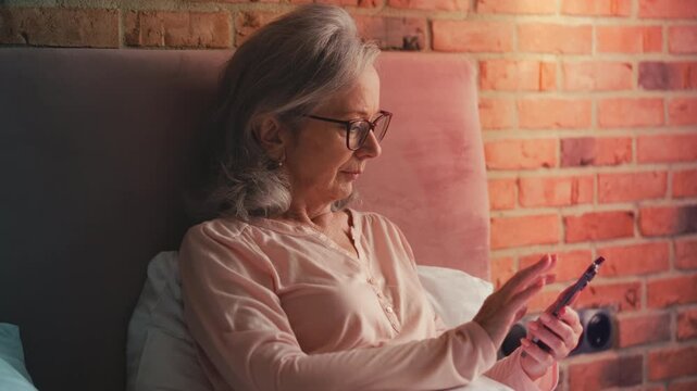 Relaxed senior woman scrolling social networks on phone, resting in bed
