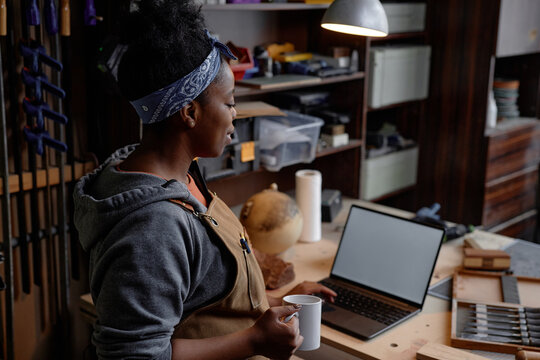 Woman in workshop holding cup while working on laptop, surrounded by tools and materials on workbench, demonstrating focus and creativity, wearing headband