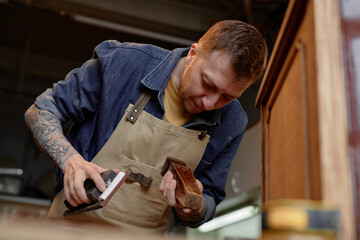 Woodworker concentrating on sanding wooden furniture piece, wearing apron and surrounded by workshop tools and equipment. Tattoed arms visible while engaged in meticulous craftsmanship