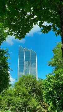 A modern high-rise glass building surrounded by trees. The building consists of three distinct sections with a sloping roof and reflective glass surfaces reflecting the blue sky and surrounding enviro