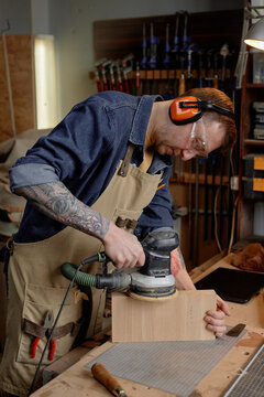 Man using electric sander on wooden block in workshop while wearing protective gear and seen focused on his task. Background features tools and equipment for woodworking projects