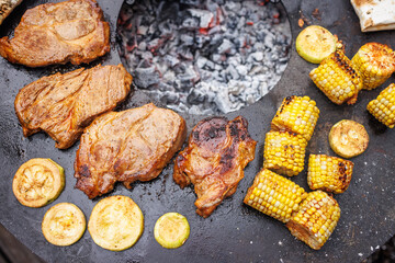 round grill with meat steaks, fish, corn, and zucchini, top view against garden background
