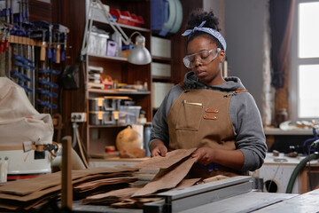 Naklejka premium Woman working in woodworking workshop, wearing protective goggles and apron, holding leather pieces. Background filled with crafting tools, shelves with supplies