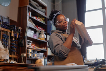 African American woman working on wooden piece in workshop, wearing protective eyewear and apron while standing next to window. Shelves filled with various tools and supplies in background