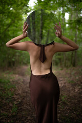 A woman holding a round mirror that reflects the trees, blending with the forest. Mode with tight brown dress standing back to camera. Image contains noise and motion blur. Selective focus