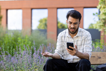 Worried and frustrated young indian man wearing glasses sitting outside on a bench and looking upset at the mobile phone screen while waving his hand incoherently