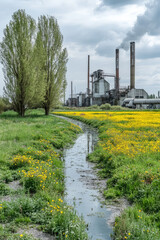Obraz premium Green flowering field with trees and an industrial area with a factory, pipes and desolation