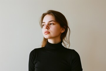 Obraz premium A young woman in a black turtleneck stands against a plain backdrop, her gaze directed thoughtfully to the side, exuding a contemplative mood.