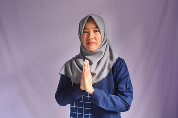 Portrait of smiling beautiful young Asian Muslim woman wearing hijab and white shirt standing with Eid greeting gesture and welcoming Ramadan isolated on orange studio background