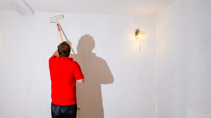 A Caucasian man painting a white wall with a roller to revamp and refresh his home during a DIY...