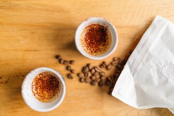 Top view of two cups of coffee with scattered coffee beans and a white coffee bag on a wooden table