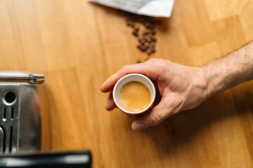 Top view of a hand holding a cup of freshly brewed espresso next to an espresso machine