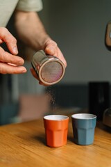 Close-up of a person sprinkling cocoa powder on two espresso cups on a wooden surface.