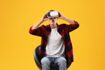Emotional young man with virtual reality headset sitting on chair against yellow background