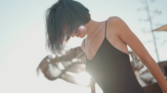 Carefree young woman with short black hair and black dress dances by the beach at sunset.