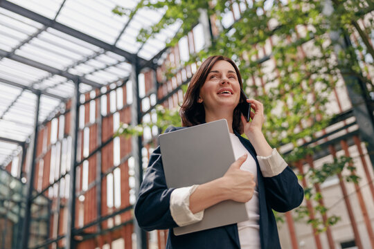 A successful businesswoman in a modern glass building holding a laptop while on a phone call - Powered by Adobe