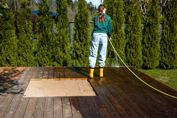 Young woman cleaning a wooden deck terrace with a hose in a sunny backyard