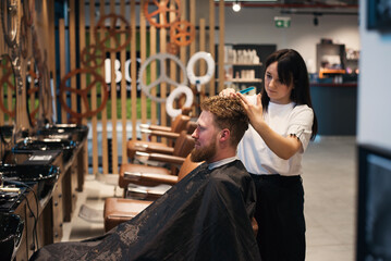 Red-haired bearded man sitting in barbershop and female barber making hairdo to guest