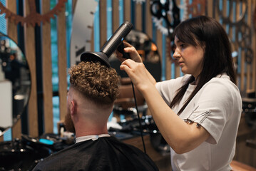 Professional woman-hairdresser is drying the hair of a curly red-haired man with hairdryer.