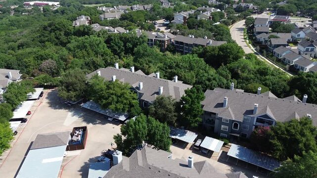 Park side apartment complex surrounding lush green trees, large dumpster, covered parking structure in suburbs Dallas Fort Worth metroplex, multistory condos townhome buildings shingle roofing