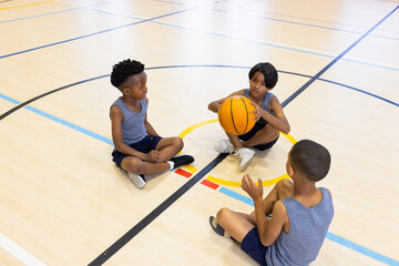 In school gym, three kids sitting on floor holding basketball, talking
