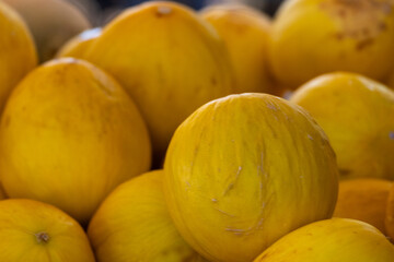 A bin of elongated vibrant yellow canary melons. The rind or skin is hard and corrugated with a waxy surface. The bulk supply is for sale at a market. The edible melons are a firm and sweet fruit.