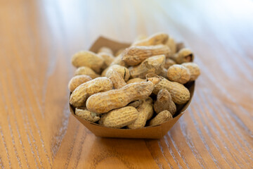 A paper basket containing dried salted peanuts on a wooden table. The salty snacks are whole with the shells dried. The tubular-shaped protein is tan-colored with a papery texture on the outside nut.