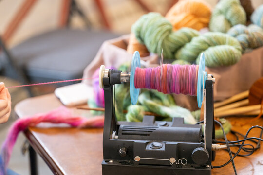 A woman uses an electric spinning wheel with colorful pink wool the handspun textured yarn on a wooden table. There's a box of balls of green yarn in the background. The wheel is blue colored plastic.