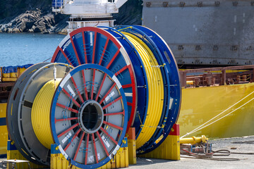 Bulk sub-sea industrial glass fiber optic cable on a metal spool on a ship's stand. The yellow data line is coiled around a black reel in a storage yard. Internet communications spool storage yard.