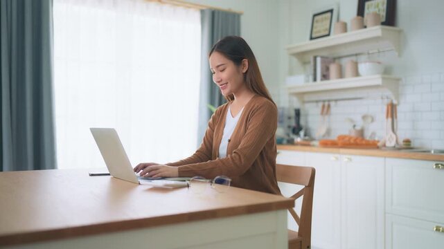 Beautiful young Asian woman working with laptop computer in home kitchen. Work at home