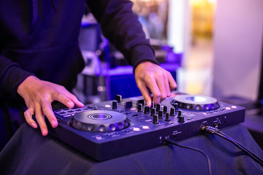 Close-up of a DJ's hands operating a modern digital DJ controller with illuminated blue and orange buttons, in a dark nightclub setting with atmospheric purple lighting.