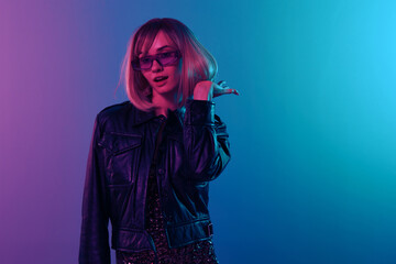 Sexy beautiful blonde woman in leather jacket sparkly dress trendy sunglasses touch hair look aside posing isolated in color light studio wall background. Neon party Cyberpunk concept. Copy space