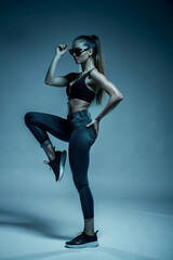 Full body side view of young fit woman with ponytail in street style outfit and sunglasses posing in studio with raised leg
